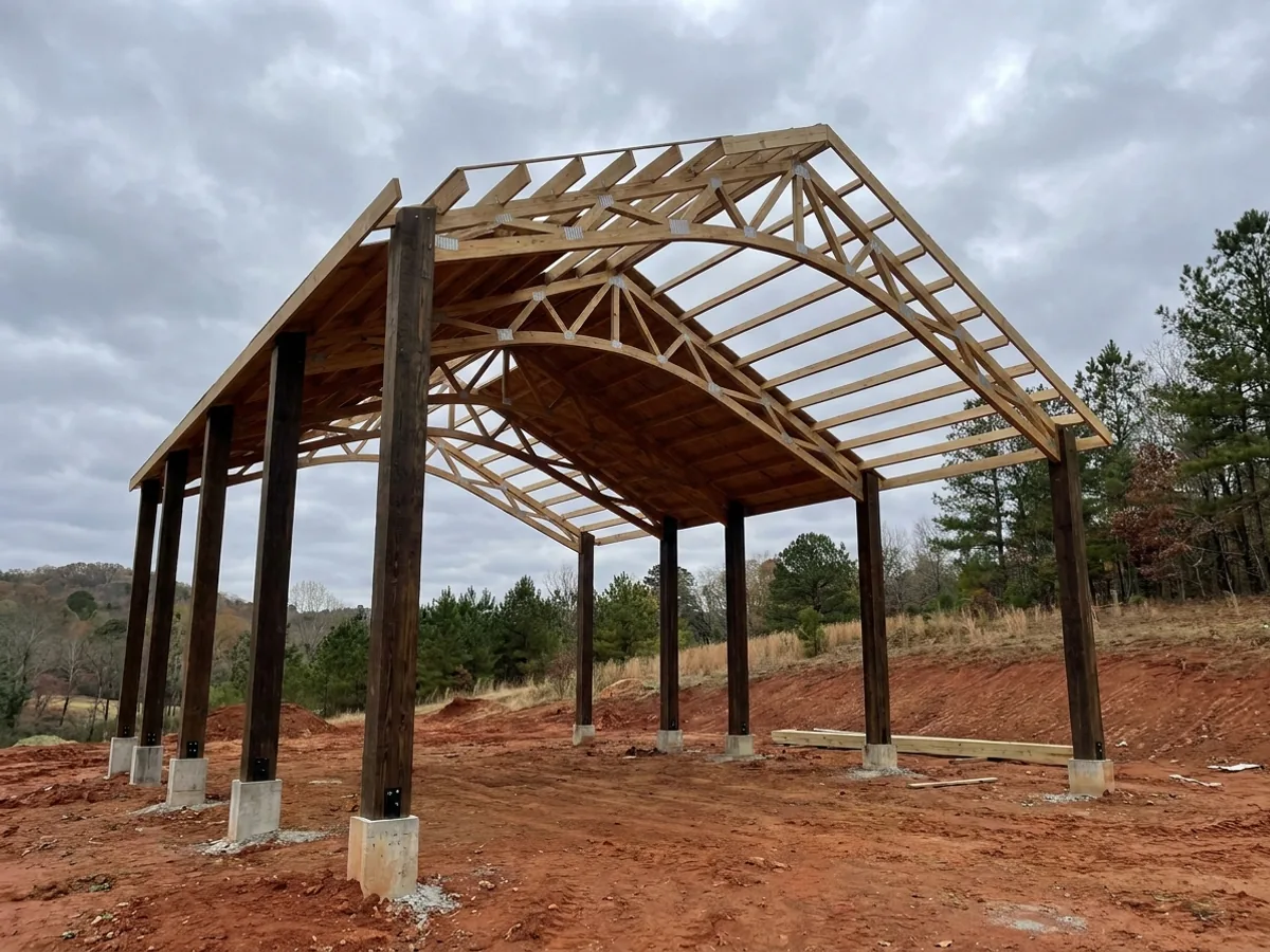 A post-frame home under construction, showing laminated timber columns on concrete piers and wide-span roof trusses with no interior bearing walls.